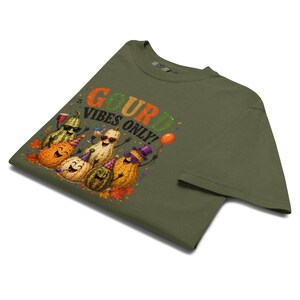 Op de afbeelding: Een olijfgroen t-shirt met een grafische print van vijf antropomorfe kalebassen die feestmutsen en zonnebrillen dragen. De tekst "GOURD VIBES ONLY!" staat boven de kalebassen, die omringd zijn door herfstbladeren en confetti.