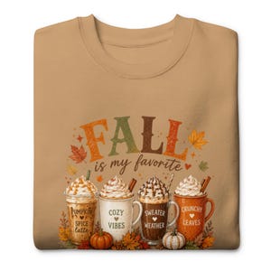 May include: A tan crewneck sweatshirt featuring a graphic print with the text FALL is my favourite. The design includes four mugs of coffee topped with whipped cream, labelled Pumpkin Spice latte, Cozy Vibes, Sweater Weather, and Crunchy Leaves.