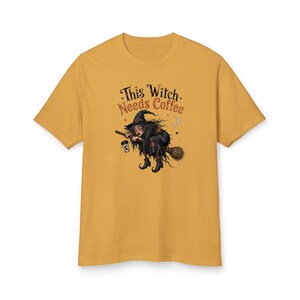 Può includere: Una t-shirt a maniche corte color giallo senape con la grafica di una strega che vola su una scopa tenendo in mano una tazza di caffè. Il testo sopra la strega recita "This Witch Needs Coffee" in un carattere stilizzato, con stelle e una luna.