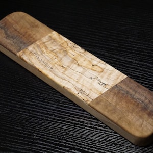 May include: A rectangular wooden board featuring a central section of light-colored, figured wood with intricate grain patterns, flanked by two darker, solid brown wood end pieces. The board has rounded corners and a smooth, polished finish.