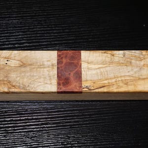 May include: A rectangular wooden case featuring a light tan, swirling wood grain pattern on the left and right sides, separated by a central vertical band of dark reddish-brown burl wood with a distinct, textured, circular pattern.