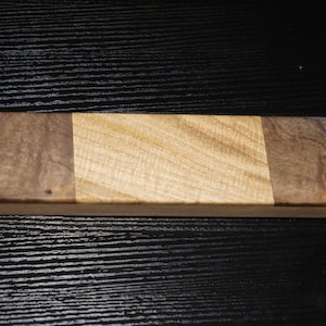 May include: A rectangular wooden case with rounded corners, featuring a light-colored, textured wood center panel flanked by two darker, marbled wood sections on either side, creating a three-part horizontal design across the top surface.