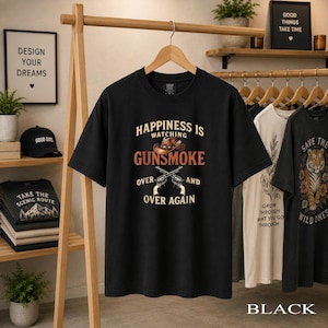 May include: A black short-sleeved t-shirt featuring a graphic print with the text "HAPPINESS IS WATCHING GUNSMOKE OVER AND OVER AGAIN". The design includes a brown cowboy hat and two crossed revolvers in a vintage-inspired colour palette.