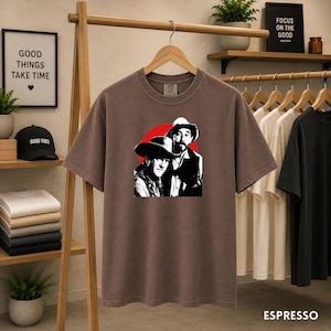 May include: A brown short-sleeve t-shirt featuring a graphic print of two individuals wearing cowboy hats against a red circular background. The shirt has a crew neckline and is displayed on a wooden hanger. The word ESPESSO is printed at the bottom.