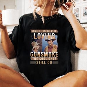 May include: A black short-sleeved t-shirt featuring a graphic print of two men in cowboy hats from the television show Gunsmoke. The text reads: SOME OF US GREW UP LOVING GUNSMOKE THE COOL ONES STILL DO. The design includes tan and white lettering.