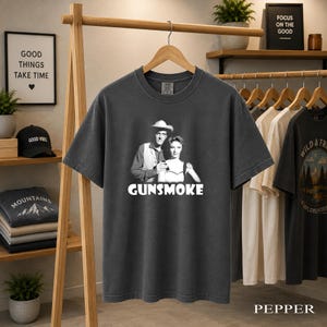 May include: A charcoal grey short-sleeved t-shirt featuring a black and white graphic of a man in a cowboy hat and a woman standing together. The word GUNSMOKE is printed in bold white capital letters below the figures on the front of the shirt.