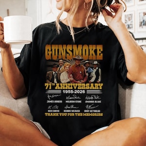 May include: A black t-shirt featuring a graphic print of the Gunsmoke 71st Anniversary 1955-2026. The design includes a cast photo, the show title in gold, and the printed signatures and names of James Arness, Milburn Stone, Amanda Blake, Ken Curtis, Dennis Weaver, and Burt Reynolds, with the text Thank You For The Memories.