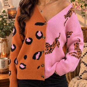 May include: A long-sleeve button-down cardigan featuring a split design. The left side is a burnt orange color with black and pink leopard spots, while the right side is light pink with a repeating pattern of small orange and black tigers.