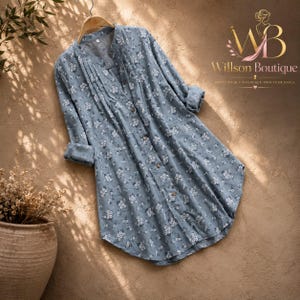 Op de afbeelding: Een tuniek met lange mouwen en knoopsluiting in een stoffige blauwe kleur met een kleine witte bloemenprint. Het kledingstuk heeft een opstaande kraag, verticale plooien langs de knoopsluiting aan de voorzijde en omgeslagen mouwen. Tekst Willson Boutique.