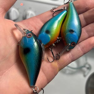 May include: Three fishing lures with a teardrop shape, featuring a gradient color scheme of metallic blue, green, and orange. Each lure has a clear plastic diving lip, a black spot on the side, red-rimmed eyes, and silver metal treble hooks.