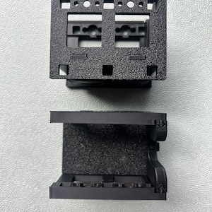 Peut inclure: Deux composants en plastique noir imprimés en 3D avec une finition texturée. La pièce supérieure présente un cadre rectangulaire avec plusieurs découpes carrées et circulaires. La pièce inférieure est un support en U avec des renforts latéraux.