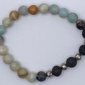 Könnte beinhalten: Ein Perlenarmband mit einer kreisförmigen Anordnung aus glatten, runden, mehrfarbigen Steinperlen in Hellblau, Creme und Beige, die in einen Abschnitt aus dunklen, porösen Lavaperlen übergehen, welche mit kleinen, silberfarbenen Zwischenelementen versehen sind.