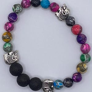 May include: A stretch bracelet featuring a variety of multi-colored, marbled round beads in shades of pink, purple, blue, green, yellow, and black. The design includes three silver-toned metal elephant charms and three porous black lava beads.