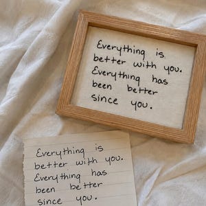 May include: A light wood frame containing white fabric embroidered with black text that reads: Everything is better with you. Everything has been better since you. Beside it lies a piece of white paper with the same text written in black ink.