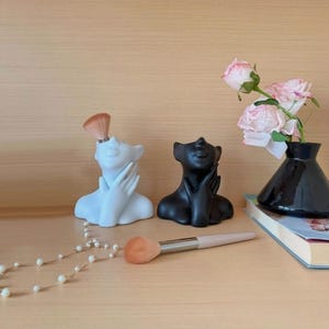 May include: Two head-shaped organizers in matte white and black, each with a hand touching the chin. A makeup brush is inserted into the white organizer, while another lies horizontally. A black vase holds pink roses next to a pearl necklace.