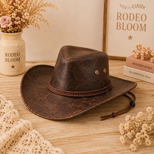 Op de afbeelding: Een donkerbruine cowboyhoed van leer met een verweerde uitstraling, voorzien van een ingedeukte kroon, een brede rand en een decoratieve gevlochten koordband. Aan de zijkant zitten twee kleine metalen klinknagels en een dunne kinband.
