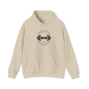 Könnte beinhalten: Ein beigefarbener Kapuzenpullover mit einer zentralen Grafik einer schwarzen Hantel mit einem Kreuz in der Mitte, umgeben vom Text FAITH N FIT LIFE und den Wörtern FAITH, HEALTH, PURPOSE am unteren Rand, inklusive eines kleinen Herzsymbols.