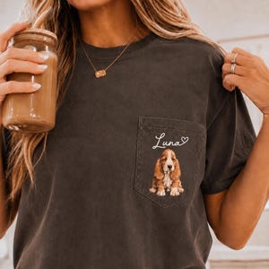 May include: A charcoal gray short-sleeve t-shirt featuring a chest pocket. The pocket displays a graphic of a brown and white Basset Hound dog sitting down, with the name Luna written in white cursive script above a small white heart outline.