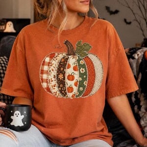 May include: A burnt orange short-sleeve t-shirt featuring a large central pumpkin graphic. The pumpkin is composed of vertical patchwork segments in various patterns, including red plaid, floral prints, and green foliage, with a brown stem.