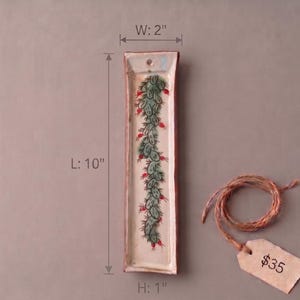 May include: A rectangular ceramic tray with a cream-colored base featuring a hand-painted green cactus design with red accents. The tray includes a small hole at the top for hanging and measures 10 inches in length, 2 inches in width, and 1 inch in height.
