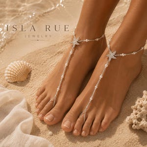 Peut inclure: Une paire de sandales pieds nus argentées avec une breloque étoile de mer incrustée de cristaux clairs, reliée à une fine chaîne ornée de petites perles blanches qui entoure la cheville et le deuxième orteil. Le texte indique ISLA RUE JEWELRY.