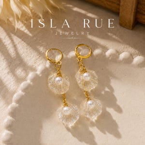 May include: Gold-tone hoop earrings featuring two dangling clear, textured shell-shaped charms and two white spherical pearls per earring. The text ISLA RUE JEWELRY is visible at the top of the image. The earrings have a hinged clasp closure.