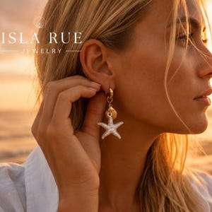 Puede incluir: Un pendiente de aro dorado con un conjunto de dijes colgantes que consiste en una pequeña concha marina dorada y una estrella de mar blanca texturizada. La joya lleva el texto ISLA RUE JEWELRY en la esquina superior izquierda.