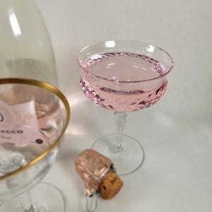 May include: A pink sparkling wine in a crystal coupe glass with a gold rim. The bottle of wine is in the background with a gold rim and a cork on the table.