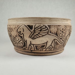 May include: A ceramic planter with a brown and white design featuring a winged lion. The planter has a textured surface and a rounded shape.