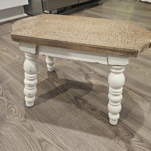 Puede incluir: Un taburete pequeño de madera con una superficie rectangular de acabado texturizado en color marrón claro y cuatro patas blancas torneadas. El taburete presenta un aspecto desgastado con desgaste visible de pintura en las patas y bordes.