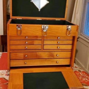 May include: A wooden tool chest with a natural finish, featuring eight drawers with silver-toned knobs, three metal latches on the front, and a hinged lid with an interior diamond-shaped mirror. The interior is lined with dark green felt.