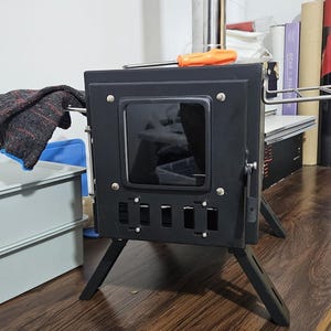 May include: A portable black metal wood stove featuring a square glass viewing window, a vertical chimney pipe, and four angled legs. An orange-handled screwdriver rests on the top surface, and a dark textured cloth hangs from the side handle.
