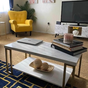 May include: A rectangular coffee table with a white faux marble top and a lower shelf, supported by four gold-toned metal legs. The table holds a silver laptop, a stack of three hardcover books, and a gold decorative tray with two gold spheres.