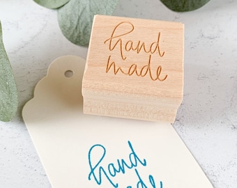 May include: A wooden stamp with the words "hand made" in a cursive font. The stamp is on a white surface with a tag that has the same words stamped in blue ink.