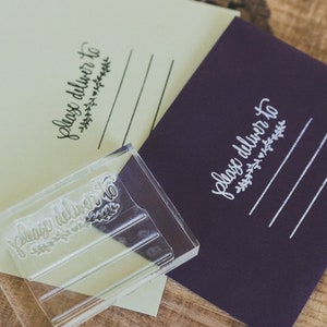 May include: A clear acrylic stamp with the text "Please deliver to" and a small floral design. The stamp is on top of a purple envelope with the same text and design stamped in white.
