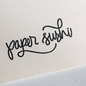 May include: Handwritten calligraphy on a white background that says "paper sushi".