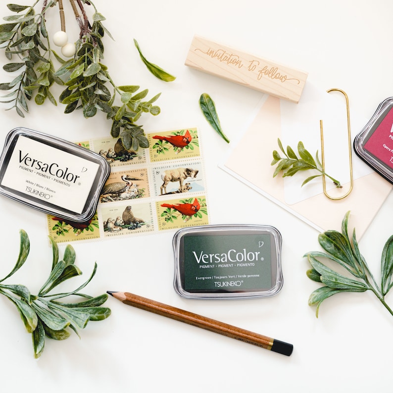 K&ouml;nnte beinhalten: Zwei gr&uuml;ne Stempelkissen mit dem Text "VersaColor" und "TSUKINEKO" darauf. Die Stempelkissen sind umgeben von gr&uuml;nem Laub, einem Holzstempel mit dem Text "invitation to follow" und einer Sammlung von alten Briefmarken.