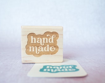 Könnte beinhalten: Ein Holzstempel mit den Worten "hand made" in einer dekorativen Schriftart. Der Stempel ist hellbraun und liegt neben einem passenden Aufkleber mit dem gleichen Text in Türkis. Der Hintergrund ist ein weiches, verschwommenes Rosa und Weiß.
