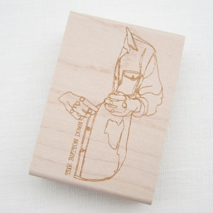 May include: A wooden stamp featuring a line drawing of a person fastening a shirt. The text "THE BUTTON DOWN" is printed on the stamp.