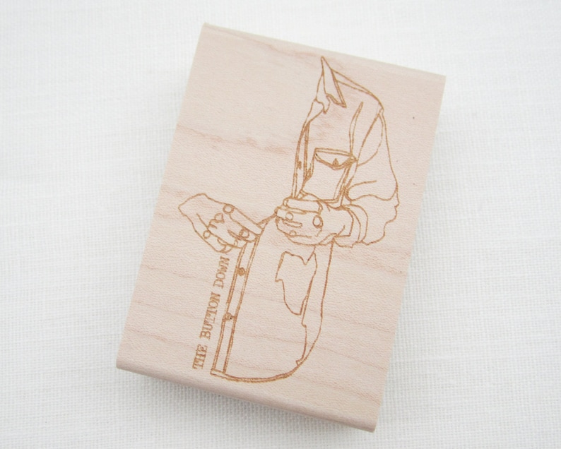 May include: A wooden stamp featuring a line drawing of a person fastening a shirt. The text "THE BUTTON DOWN" is printed on the stamp.