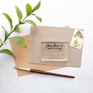 May include: A clear rubber stamp with the text "Please deliver to" and a decorative floral design. The stamp is on a beige envelope with two postage stamps.