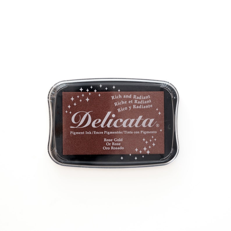 May include: A brown ink pad with the text "Delicata" in a white script font. The ink pad is labeled "Rich and Radiant, Riche et Radiant, Rico y Radiante" and "Rose Gold, Or Rose, Oro Rosado".