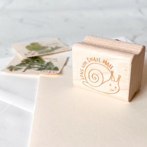 May include: A wooden stamp with the words "LONG LIVE SNAIL MAIL" and a snail illustration. The stamp is next to several postage stamps with tree illustrations and blank stationery.