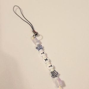 May include: A phone charm featuring a black cord, two translucent light purple rectangular beads, two glittery blue star-shaped beads, and three white round beads with the black letters T, X, and T printed on them, arranged in a vertical sequence.