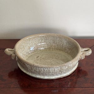 May include: A round ceramic baking dish with a speckled beige and cream glaze. The dish features two small, looped handles on opposite sides and a wide, shallow basin with a smooth, glossy finish throughout the interior and exterior surfaces.