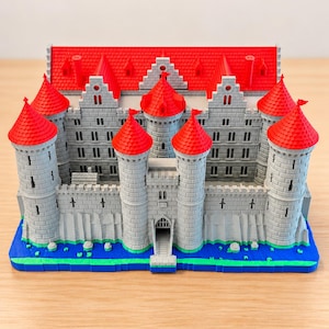May include: A 3D printed model of a castle featuring grey stone-textured walls, bright red conical roofs, and a central gatehouse. The structure sits on a blue and green base representing water and land, with multiple towers and windows.