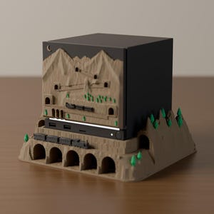 May include: A black rectangular electronic device dock encased in a tan, 3D-printed mountain landscape. The base features a multi-arched stone bridge with a small black train, while the upper section depicts carved mountain peaks and tunnels.