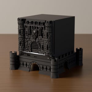 May include: A matte black, cube-shaped electronic device enclosure featuring a detailed 3D-printed facade of a medieval castle gatehouse with stone textures, towers, and a portcullis, mounted on a matching castle-themed base with corner turrets.