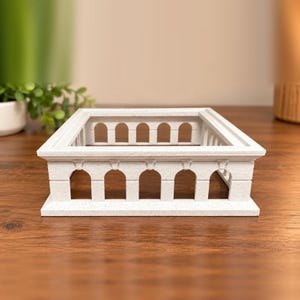 May include: A square, off-white, speckled architectural display stand featuring a series of open arches along all four sides. The structure has a flat, wide top rim and a solid base, resembling a miniature colonnade or stadium.