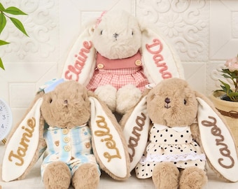 以下が含まれることがあります： 長い耳に名前が刺繍された3体のぬいぐるみウサギ。中央のウサギはピンクのギンガムチェックのドレスを着ています。左のウサギはJamieとLeilaという名前が入った青いストライプのパジャマを着ています。右のウサギは水玉模様のドレスを着ています。